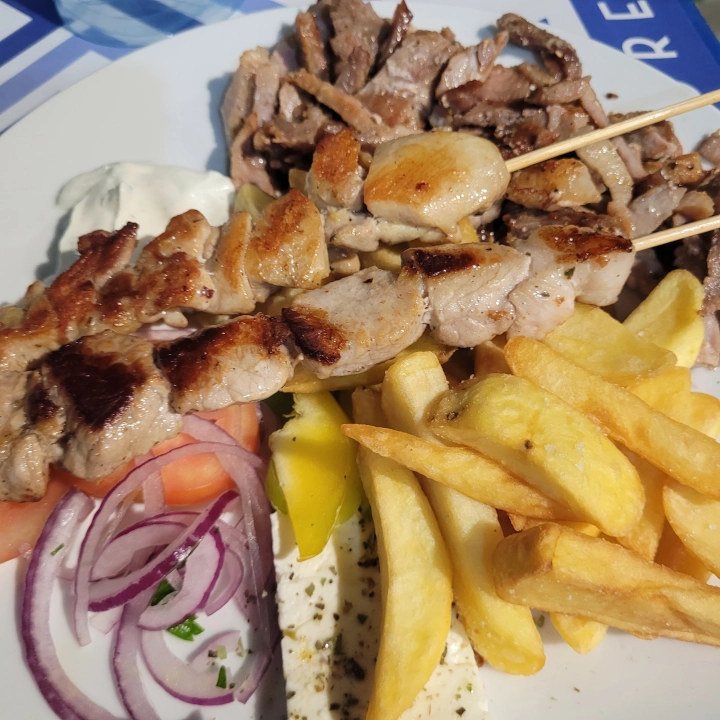 Piatto di Gyros con carne di maiale a fette, servito su una pita, accompagnato da patatine fritte, pomodoro, peperone, feta, cipolla e tzatziki